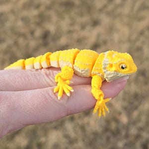 May include: A yellow, segmented, plastic toy lizard with a textured surface. The toy lizard has a small, pointed snout and a long, slender tail.