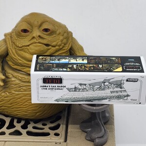 May include: A Star Wars Vintage Collection box for Jabba's Sail Barge (The Khetanna) vehicle. The box is white with black and red text and a black and white illustration of the sail barge.
