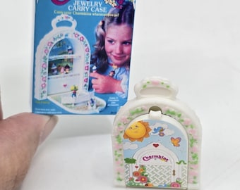 以下が含まれることがあります： 青と白の花柄の白いプラスチック製のチャームキンズジュエリーキャリーケース。ケースには、太陽、雲、蝶が描かれた蝶番付きのドアが付いています。ケースは閉まっており、ドアにはチャームキンズのロゴが見えます。
