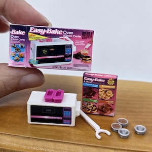 以下が含まれることがあります： ミニチュアのEasy-Bake Oven & Snack Centerおもちゃセット。小さな白とピンクのオーブン、4種類のミックスが入った箱、白いプラスチック製のツール、4つの小さな銀色の焼き型が含まれています。パッケージには画像とテキストが記載されています。