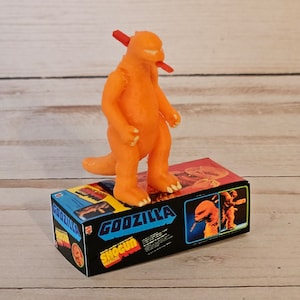 Op de afbeelding: Oranje Godzilla speelgoedfiguur met een rood zwaard in zijn mond, staand op een zwarte doos met de woorden "Godzilla Shogun Warriors" erop gedrukt.