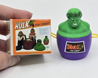 May include: A green plastic toy chest shaped like the Incredible Hulk's head. The chest has a purple lid with a sticker that says "The Incredible Hulk Toy Chest". The box shows a child playing with the toy chest.