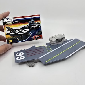 Pode incluir: Um porta-aviões de brinquedo de plástico cinza com uma pista de pouso branca e um pequeno avião de plástico cinza no convés. O porta-aviões tem o número 66 impresso no lado. A caixa do brinquedo está no fundo e diz "G.I. Joe U.S.S. Flagg Aircraft Carrier".