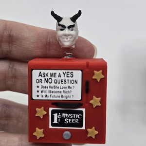 May include: A red square box with gold star accents featuring a white panel that reads ASK ME A YES OR NO QUESTION, Does He/She Love Me?, Will I Become Rich?, Is My Future Bright?, and 1¢ MYSTIC SEER. A white devil head with black horns sits on a spring.