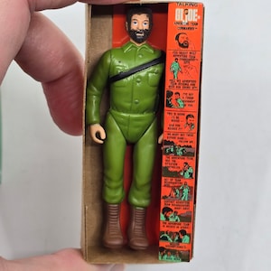 Puede incluir: Una figura de acci&oacute;n vintage de un hombre con barba que lleva un uniforme militar verde y botas marrones. La figura est&aacute; empaquetada en una caja de cart&oacute;n con fondo rojo y blanco. La caja presenta una imagen de la figura y un texto que dice "Talking Blooe Adventure Team Commander".