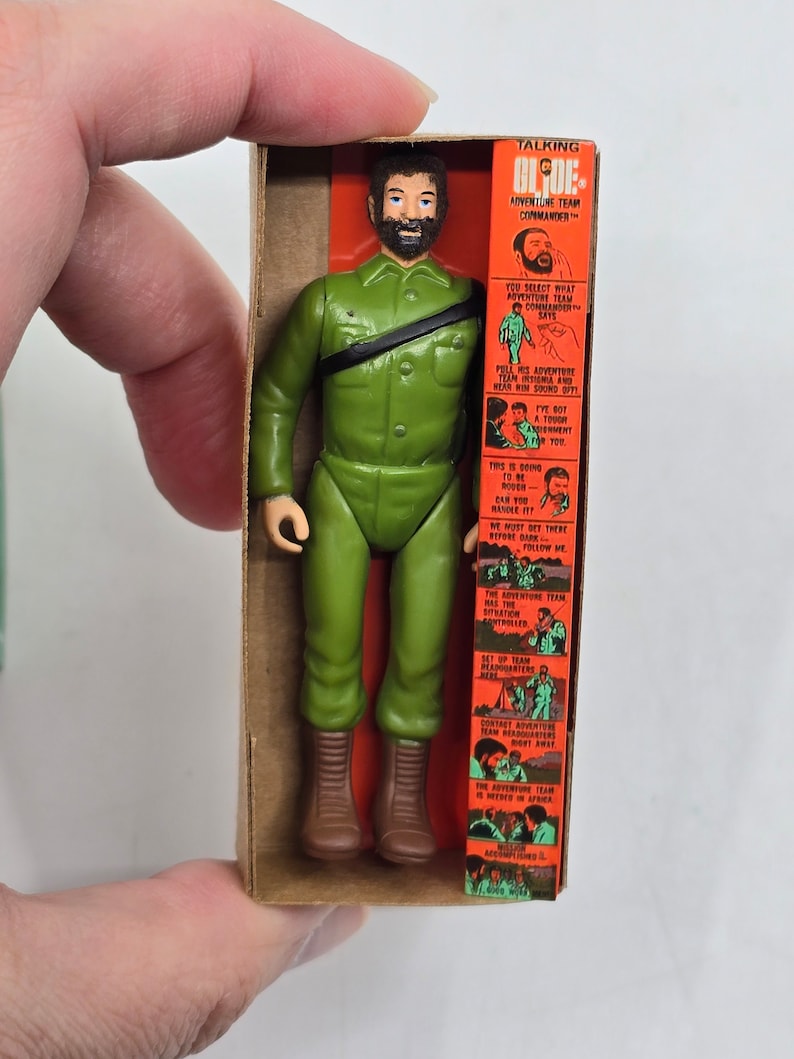 Puede incluir: Una figura de acci&oacute;n vintage de un hombre con barba que lleva un uniforme militar verde y botas marrones. La figura est&aacute; empaquetada en una caja de cart&oacute;n con fondo rojo y blanco. La caja presenta una imagen de la figura y un texto que dice "Talking Blooe Adventure Team Commander".