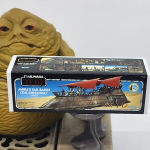 May include: A Star Wars Return of the Jedi Jabba's Sail Barge (The Khetanna) vehicle box. The box is white with blue and red accents. The box features a detailed illustration of the sail barge with Jabba the Hutt's palace in the background. The box also includes the text "Star Wars Return of the Jedi" and "Jabba's Sail Barge (The Khetanna)".