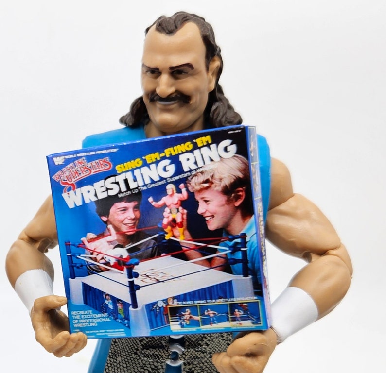 May include: A blue and white wrestling ring toy box with the text "Sing 'Em-Fling 'Em Wrestling Ring" and "Match Up The Greatest Superstars of All Time".