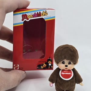 May include: A brown Monchhichi doll with a red bib and a small pink pacifier. The doll is standing next to its red and white box, which has the words "Monchhichi" and "The Original" printed on it.
