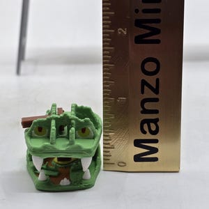 May include: A green and brown plastic toy crocodile with a hinged jaw. The crocodile is approximately 2 inches long. The toy is next to a ruler with the text "Manzo Mini" printed on it.