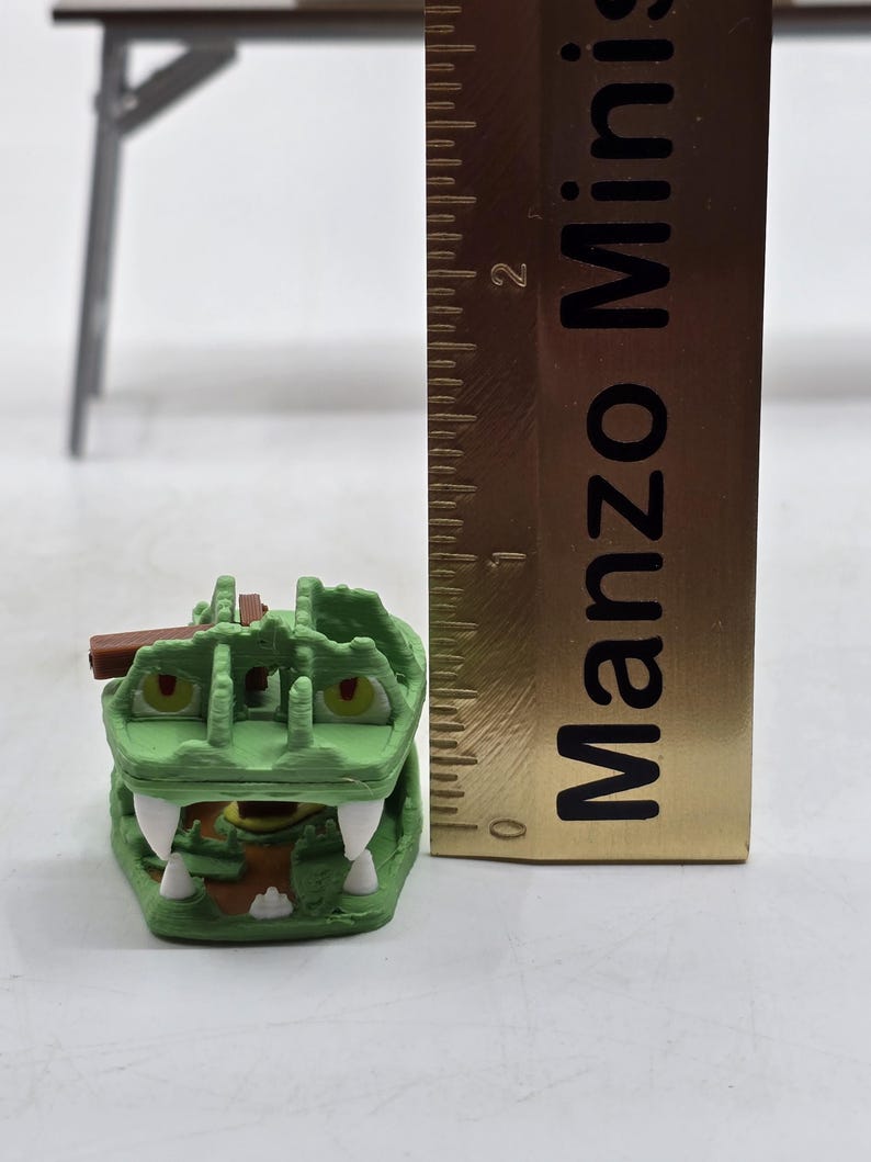 May include: A green and brown plastic toy crocodile with a hinged jaw. The crocodile is approximately 2 inches long. The toy is next to a ruler with the text "Manzo Mini" printed on it.