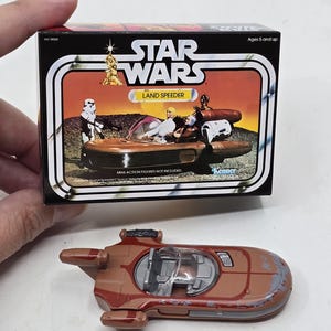 Op de afbeelding: Een vintage Star Wars Land Speeder speelgoed in bruin en grijs met een heldere voorruit. Het speelgoed zit in de originele doos, die het Star Wars-logo en de woorden "Land Speeder" heeft.