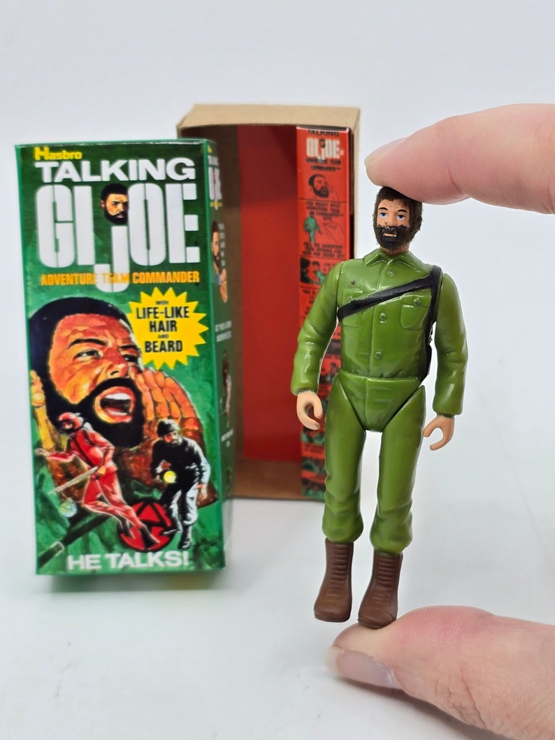 Puede incluir: Una figura de acci&oacute;n vintage Hasbro Talking G.I. Joe con uniforme militar verde, cintur&oacute;n y botas marrones. La figura sostiene un rifle y tiene pelo y barba realistas. La figura est&aacute; empaquetada en una caja con las palabras "Talking G.I. Joe Adventure Team Commander" y "He Talks!" impresas en ella.
