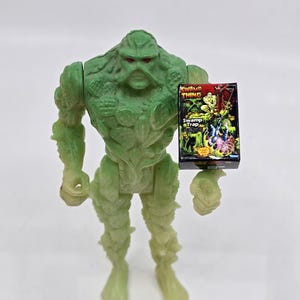 May include: A green Swamp Thing action figure holding a small box with the words "Swamp Thing" and "Swamp Trap" on it. The figure has a detailed, textured design and is posed upright. The box has a colorful illustration.