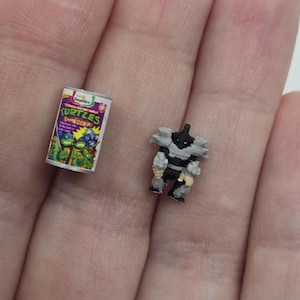 Puede incluir: Miniatura de lata de "Kurtzes Shredder" con gráficos de las Tortugas Ninja. Junto a ella, una figura de acción en miniatura de Shredder, con detalles en negro, gris y amarillo. Los artículos son pequeños y detallados.