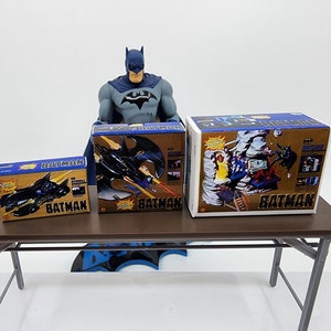 May include: Three vintage Batman toy boxes with blue and gold packaging. The boxes feature images of Batman's Batmobile and Batwing. The boxes are arranged on a brown table.