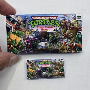 May include: A rectangular Teenage Mutant Ninja Turtles Pizza Power Game box featuring green turtles, a rat character, and city buildings. The colorful illustration shows the characters in action with the title text clearly printed on the front.