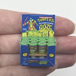 Puede incluir: Miniatura caja expositora de Teenage Mutant Ninja Turtles Retromutagen Ooze. La caja azul presenta una imagen de dibujos animados de una tortuga y el nombre del producto. La caja contiene pequeños frascos de limo con tapa verde.