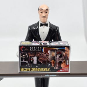 Puede incluir: Una caja para un juguete Batcave Command Center de Batman: La Serie Animada. La caja es roja, negra y blanca con imágenes de la Batcave y personajes de Batman. El texto en la caja dice "Batman: La Serie Animada" y "Batcave Command Center."