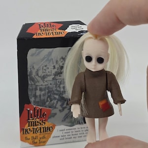 Op de afbeelding: Een kleine pop met een bruine jurk en blond haar, vastgehouden in een hand. De pop heeft grote zwarte ogen en is verpakt in een doos met de woorden "Little Miss No-Name" en "Please Take Me Home."