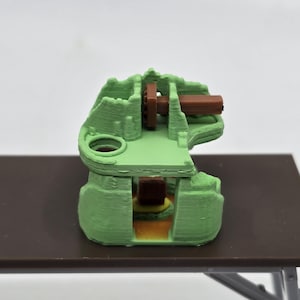 May include: A 3D printed green model of a miniature castle with a brown wooden bridge and a small brown building inside.