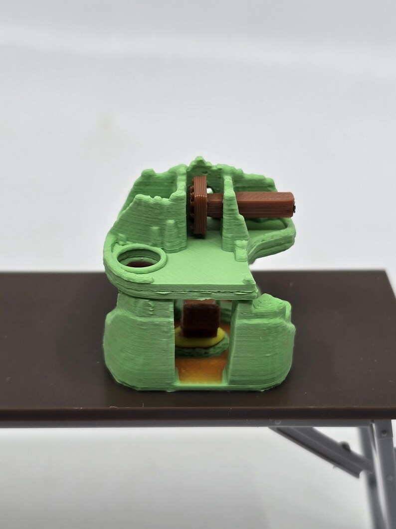 May include: A 3D printed green model of a miniature castle with a brown wooden bridge and a small brown building inside.