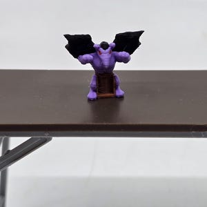 May include: Purple gargoyle figurine with black wings, standing on a brown base. The gargoyle has red eyes and a menacing expression.