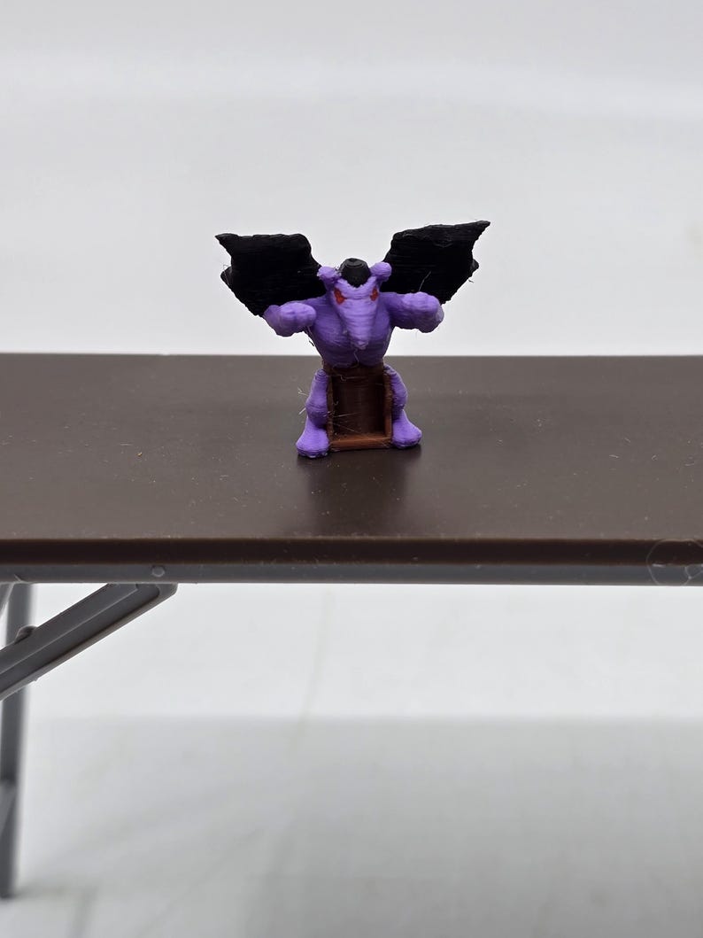 May include: Purple gargoyle figurine with black wings, standing on a brown base. The gargoyle has red eyes and a menacing expression.