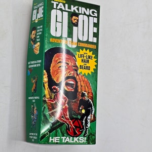 Puede incluir: Una caja verde y amarilla con una imagen de un hombre con barba y un uniforme militar verde. La caja dice "Talking G.I. Joe Adventure Team Commander" y "He Talks!" con una imagen de una placa de identificaci&oacute;n. La caja tambi&eacute;n dice "With Life-Like Hair and Beard".