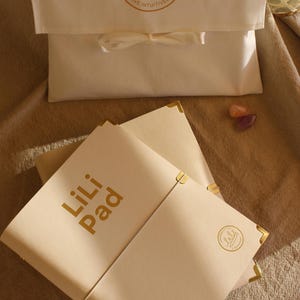 May include: Cream-coloured Lili Pad notebooks with gold corner protectors and gold lettering. A matching cream-coloured pouch with a ribbon closure and a gold logo that reads "Love Intentionally, Live Intuitively" is also in the image.