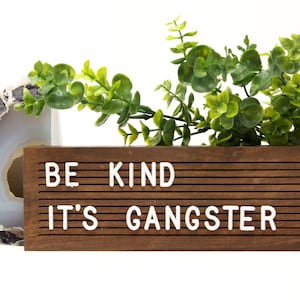 May include: A wooden letter board with the text "BE KIND IT'S GANGSTER" in white letters. The board is next to a decorative geode and a sprig of green leaves.