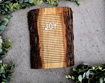 May include: A wooden letter board displaying the word "JOY" in white letters. The board features a natural wood grain and bark edges. The board is surrounded by green foliage, creating a rustic and cheerful aesthetic.