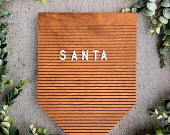May include: A wooden sign with the word "SANTA" in white letters. The sign is a warm brown colour and has horizontal lines. The sign is surrounded by green leafy branches, set against a grey background.