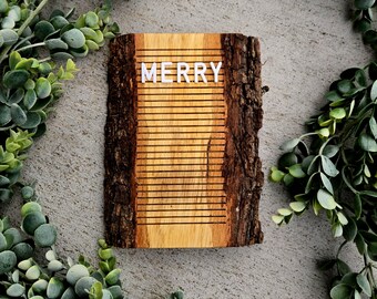 May include: A wooden sign with the word "MERRY" in white letters. The sign is made from a piece of wood with the bark still attached. The background is a grey concrete surface, and the sign is surrounded by green leafy branches.