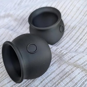 Puede incluir: Dos pequeñas ollas redondas de color negro mate con un asa circular. Las ollas tienen una abertura ancha y una base redondeada. Están sobre una superficie blanca texturizada.