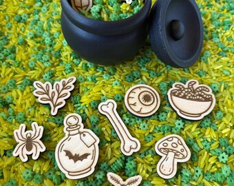 Pode incluir: Um caldeirão preto com tampa, rodeado por recortes de madeira de objetos com tema de Halloween. Os recortes incluem um cristal, uma teia de aranha, uma aranha, uma garrafa de poção com um morcego, um osso, um olho, uma tigela de maçãs, um cogumelo e uma traça. Os objetos estão sobre uma cama de arroz amarelo e verde.