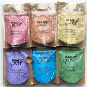 Puede incluir: Seis bolsas de "Magic Dust" en varios colores, incluyendo rosa, naranja, amarillo, morado, azul y verde. Cada bolsa está llena de purpurina y tiene la inscripción "Magic Dust". Una pequeña cuchara de madera descansa junto a la bolsa morada.