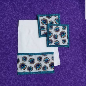 May include: Set of three white cotton pot holders with a teal border and a repeating pattern of a psychedelic eye with flames. The pot holders are on a purple background.