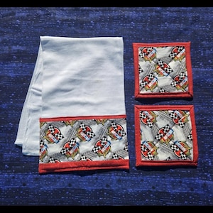 May include: A light blue kitchen towel with a patterned border featuring racing flags and logos, along with two matching square coasters. The border and coaster edges are trimmed in red.