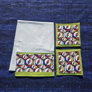 May include: A white kitchen towel and two matching square coasters with a colorful, repeating graphic pattern. The pattern features a stylized lightning bolt and skull design in red, yellow, and blue. The edges are trimmed with a green border.