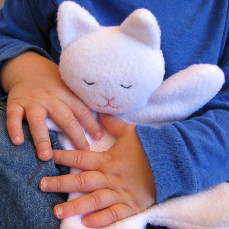May include: A white plush cat toy with closed eyes and a pink nose. The toy is being held by a child wearing a blue shirt and jeans.
