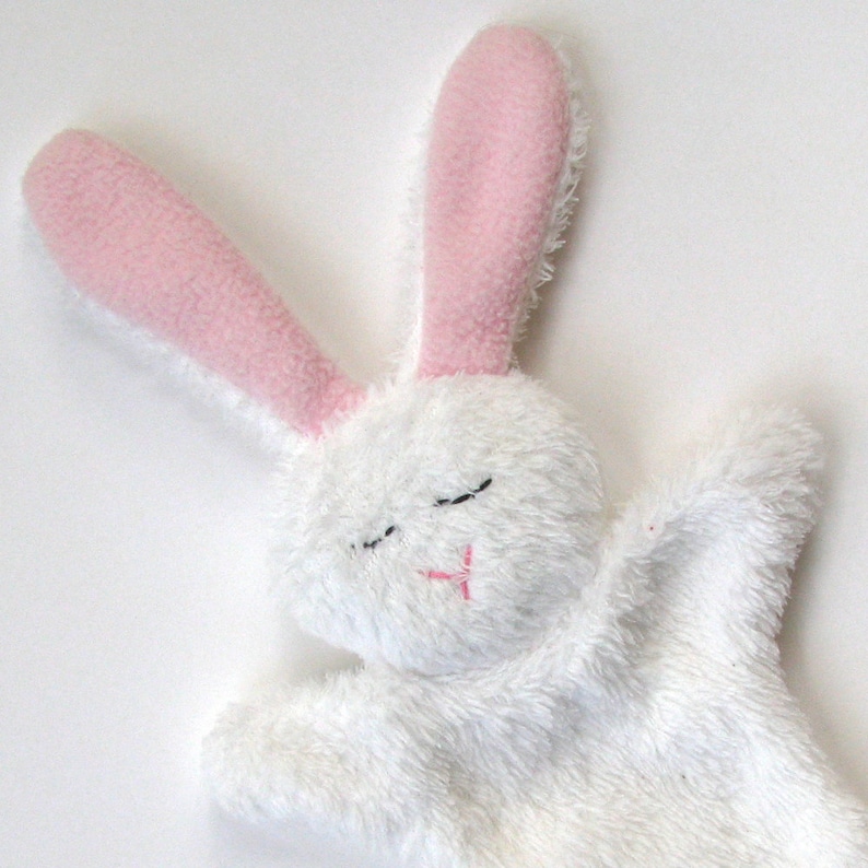 May include: A white plush bunny puppet with pink ears and embroidered eyes and mouth. The bunny is lying on its side with its arms outstretched.