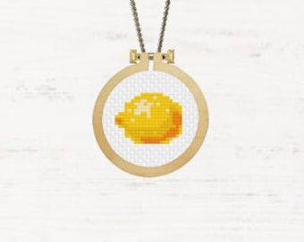May include: A cross-stitch pendant necklace featuring a yellow lemon design within a small, round wooden embroidery hoop. The pendant hangs from a delicate, bronze-colored chain. The background is a white, textured surface.