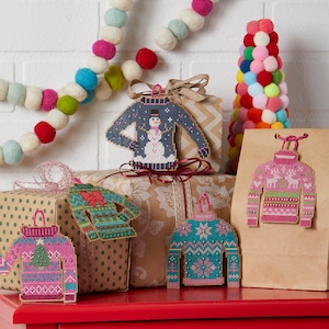 May include: Five Christmas ornaments shaped like sweaters with different patterns. The ornaments are hanging from brown gift boxes and a brown paper bag. The ornaments are decorated with snowflakes, reindeer, and a snowman.