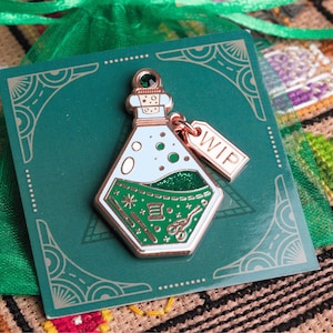 Puede incluir: Un pin de esmalte color cobre con forma de botella de poción, con un diseño verde y blanco. El pin tiene una pequeña etiqueta que dice "WIP". El pin está sobre una tarjeta verde con un borde decorativo.