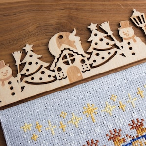 Pode incluir: Uma decoração de Natal em madeira com desenhos recortados de um boneco de neve, árvores de Natal, uma casa e candeeiros de rua. Está colocada sobre um tecido de ponto cruz azul claro com estrelas e renas bordadas a amarelo e castanho.