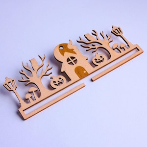 Op de afbeelding: Een lasergesneden decoratieve houten rand met een centraal huis met een maan, geflankeerd door kale bomen, pompoenen, paddenstoelen en lantaarnpalen. Het ontwerp is plat, lichtbruin en bevat een horizontale balk langs de onderrand.