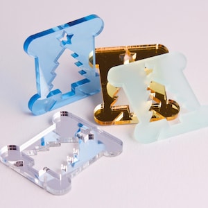Op de afbeelding: Vier kerstversieringen van acryl in blauw, goud en transparant. Elke versiering heeft een rechthoekige vorm met een uitgesneden kerstboom- en sterontwerp. De versieringen zijn gerangschikt op een wit oppervlak.