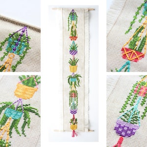May include: A vertical wall hanging with cross-stitch embroidery of hanging plants in colourful pots. The plants are green, and the pots are various colours, including purple, yellow, and red. The background is a natural linen colour, and the piece is finished with a wooden dowel and fringe.