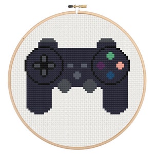 Puede incluir: Bordado de punto de cruz de un mando de videojuegos en un aro de madera. El mando es azul oscuro con botones coloridos. El fondo es blanco y el aro es de madera.